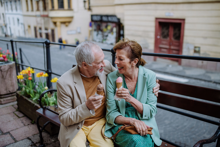 Elderly couple smiling, sitting on a bench sharing ice cream cones in a city environment