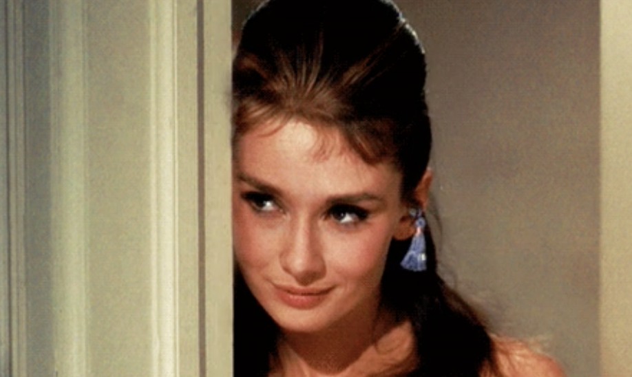 Audrey Hepburn smiles subtly, peeking around a door frame, with elegant earrings