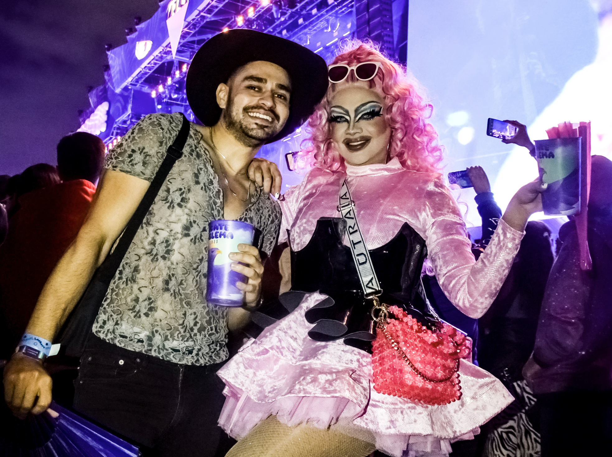 Dos personas disfrutando de un evento musical, una con sombrero y camisa estampada, la otra con atuendo rosa estilo drag queen
