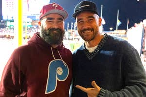 Two men smiling at a baseball game, one in a Phillies hoodie, the other pointing at him, both wearing caps