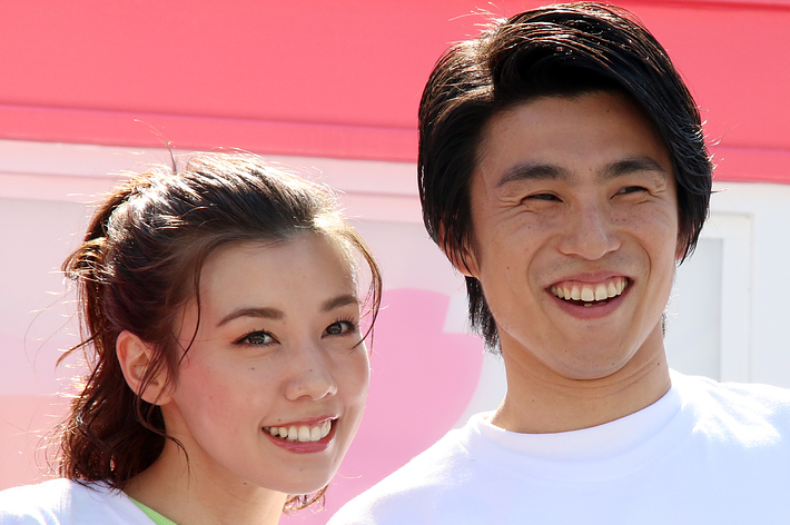 中尾明慶さんと仲里依紗さんがカメラに向かって笑顔で手を振っています。白いTシャツを着て、イベントに参加している様子です。