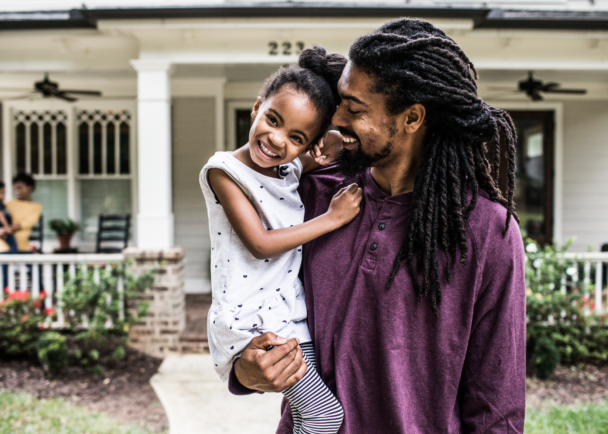 Father holding young daughter, both smiling, with a house and another person in the background