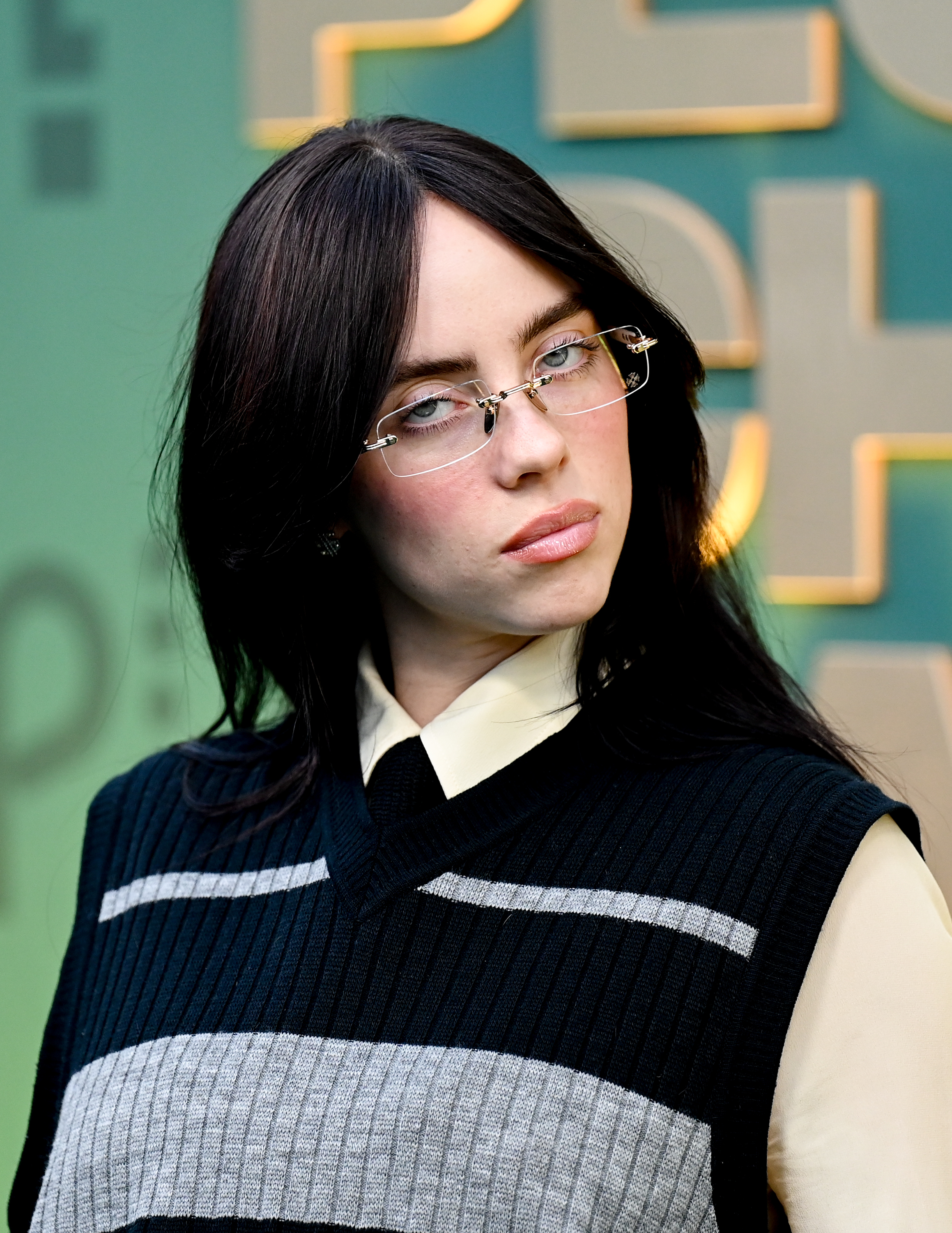 Billie Eilish in a patterned sleeveless top and glasses, posing with a slight pout