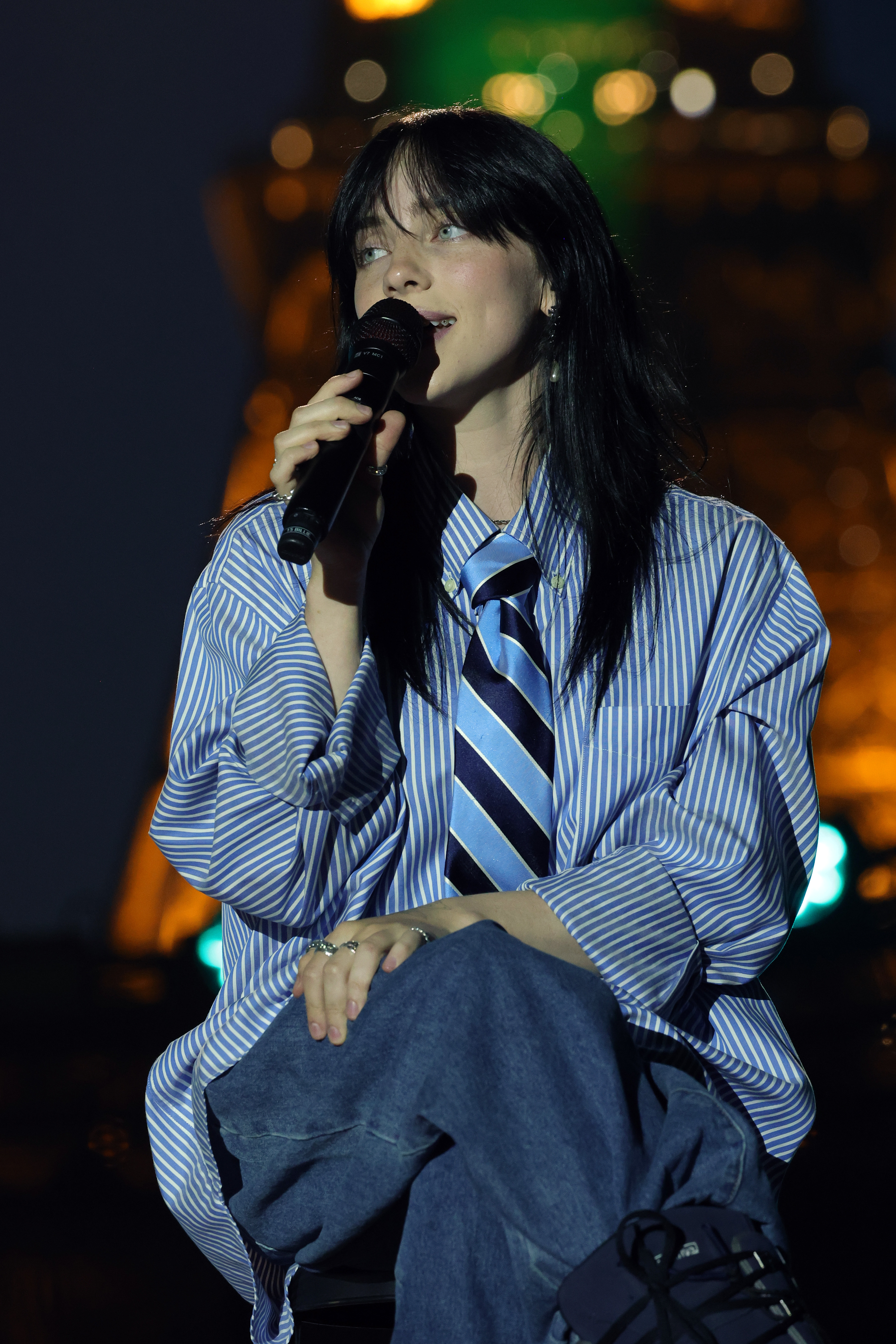 Billie Eilish performing, wearing a striped shirt with a necktie and holding a microphone
