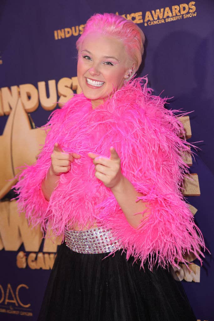 Person with pink hair and feathery top, smiling with thumbs up at an event