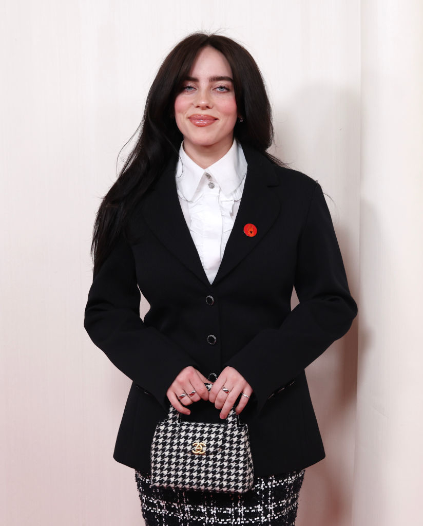 Billie Eilish standing in a blazer, shirt, with a checkered handbag and a flower pin