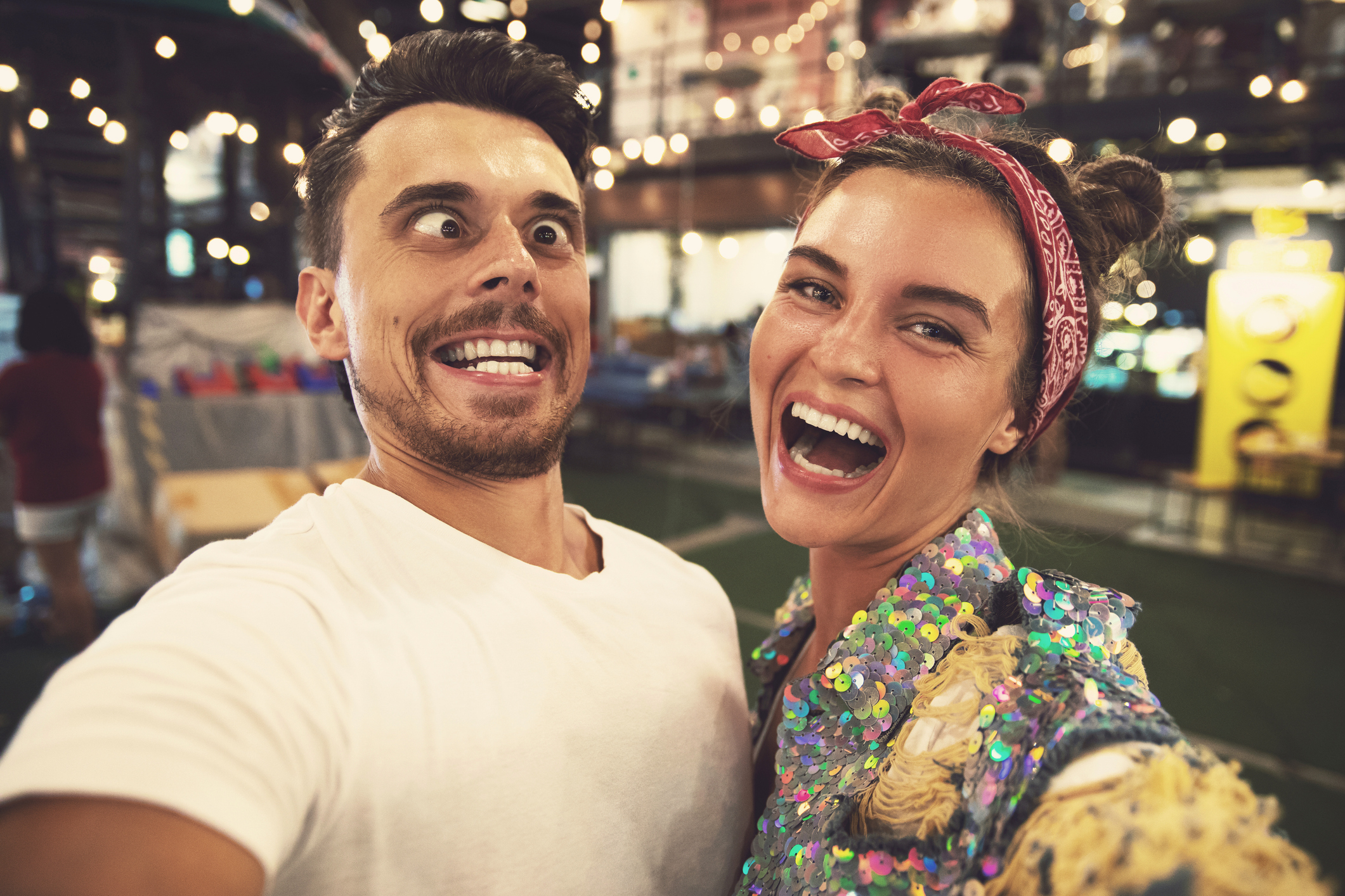 Two people taking a playful selfie, one wearing a sequined top, the other in a plain tee, both smiling widely