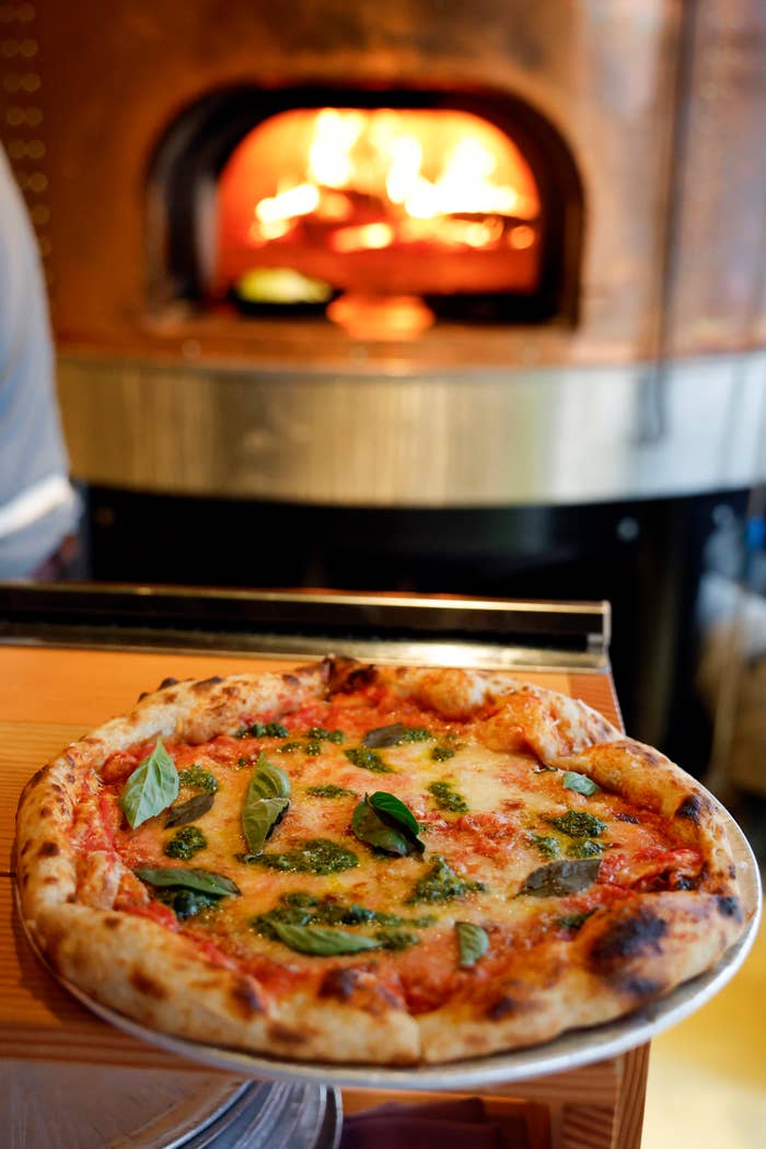 A freshly baked pizza with basil leaves in the foreground, a wood-fired oven with flames visible in the background