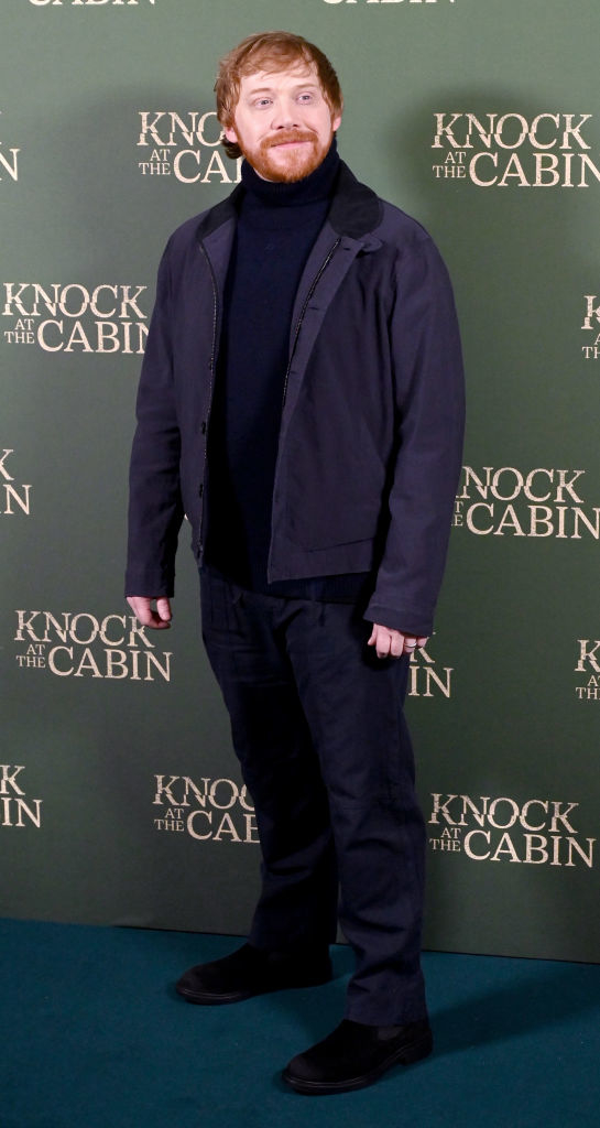Rupert in jacket and trousers with a raised eyebrow, standing before a promotional backdrop