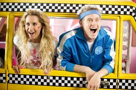 Sharpe and Ryan from High School Musical in a yellow bus with a checkered pink interior. They are smiling and posing playfully