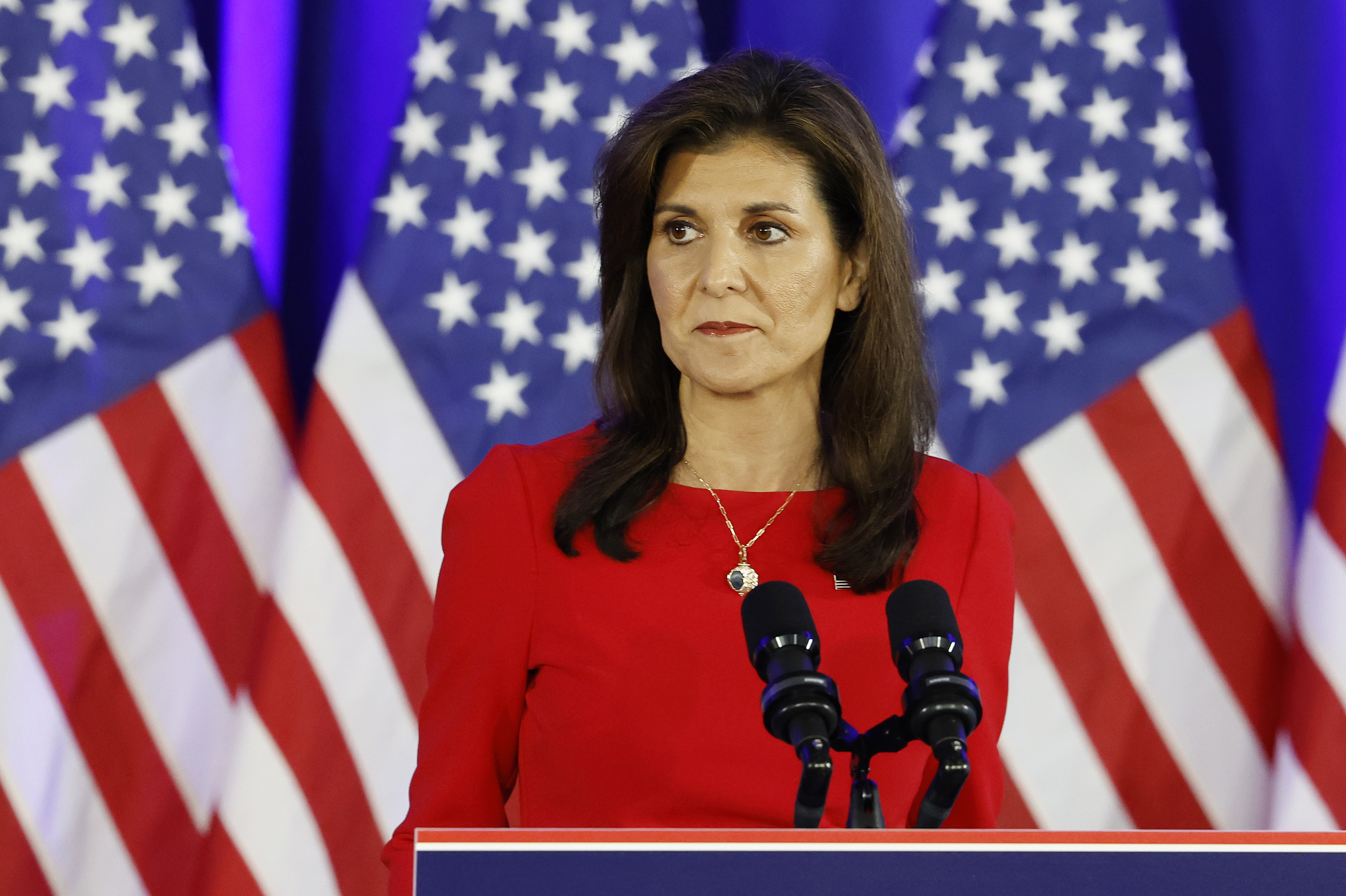 Nikki stands at a podium with multiple American flags in the background, wearing a red top and a necklace