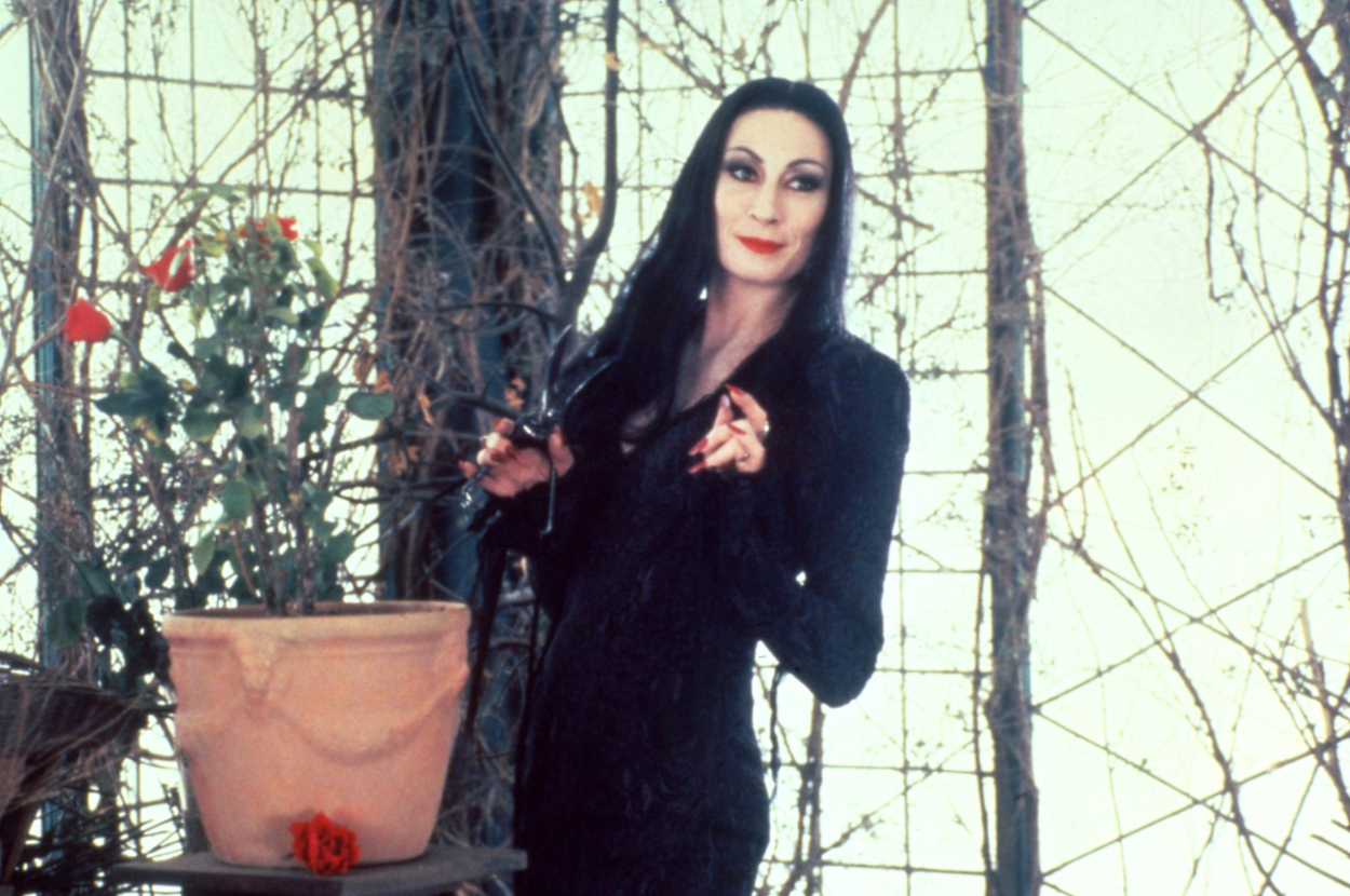 Morticia Addams stands elegantly with shears near a rose bush, dressed in her classic long black gown