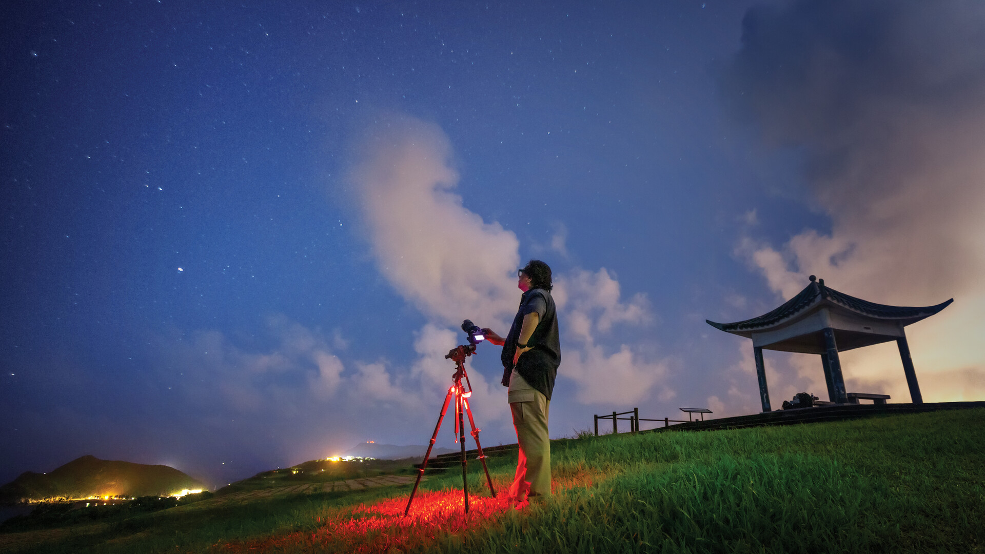 Person with a camera on a tripod under a starry sky, standing near a traditional structure