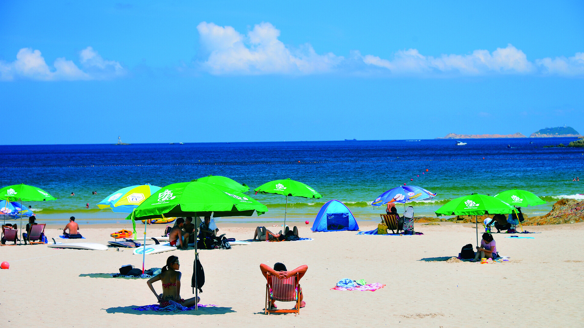 Beach scene with visitors under umbrellas, some lounging on chairs, with ocean backdrop