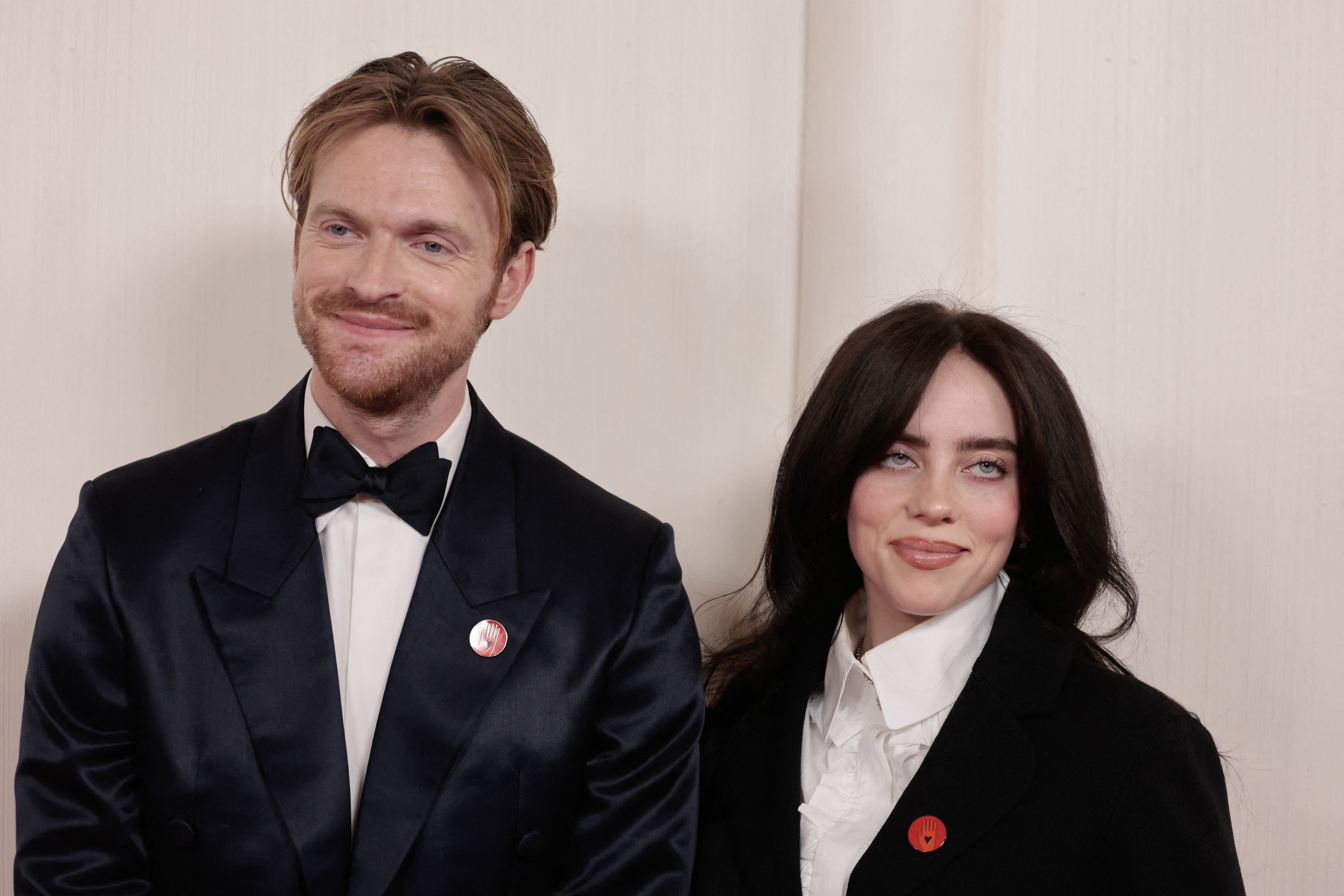 Finneas and Billie Eilish standing side by side, one in a black tuxedo and the other in a black and white suit, both wearing red lapel pins