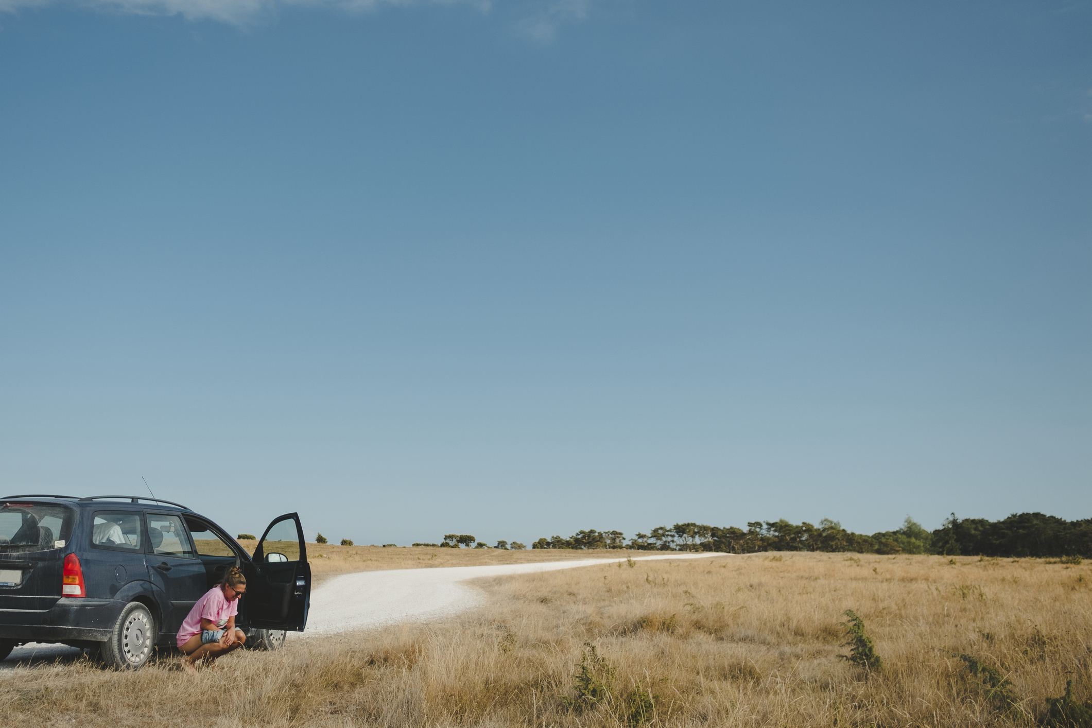 Person sitting by car on dirt road in open field, clear sky, concept of travel and leisure