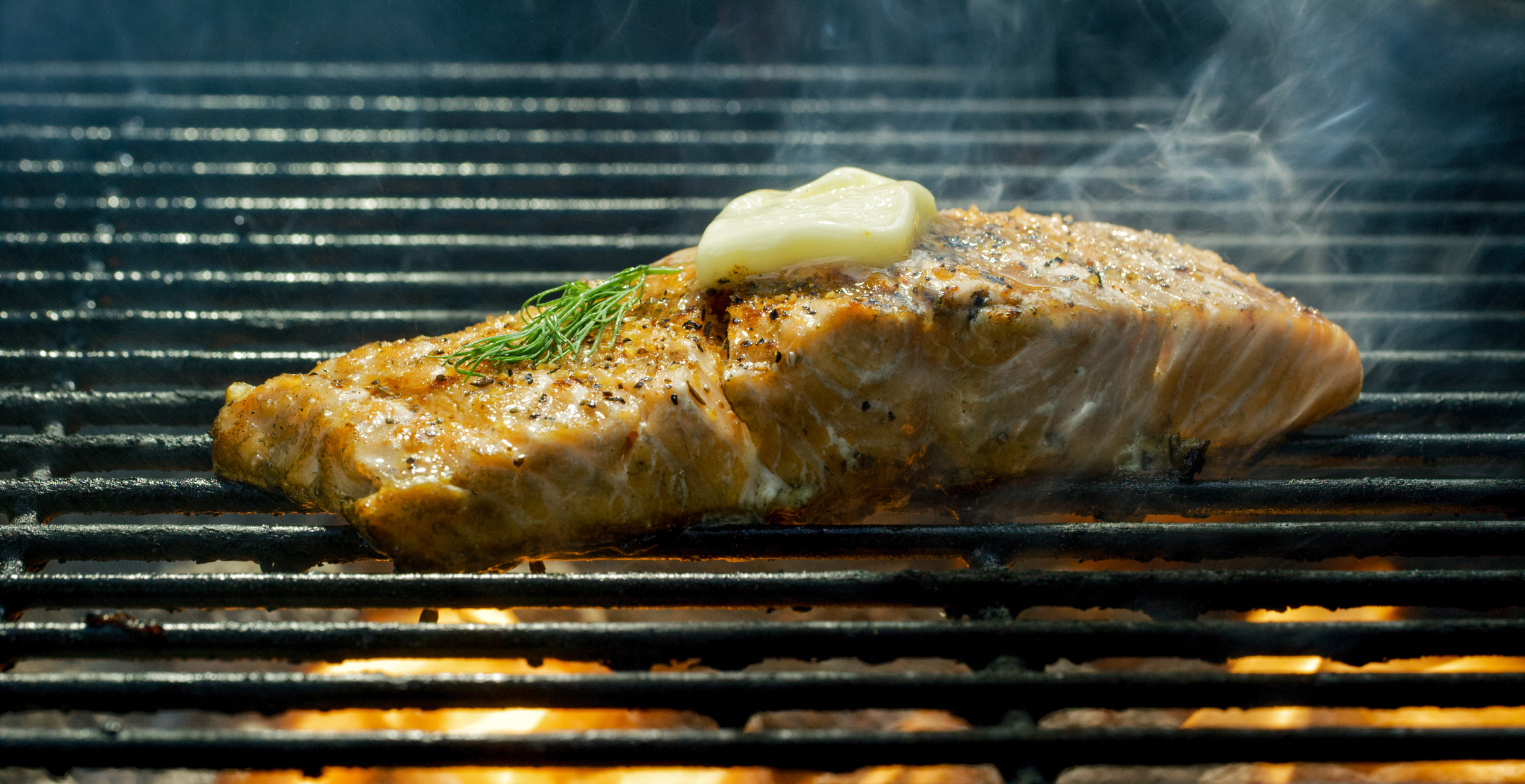 Grilled salmon with a slice of butter and dill on top, steam rising, cooking on a grill
