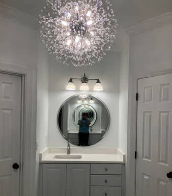Person taking a photo in a mirror in a well-lit bathroom with a chandelier and sconce lights