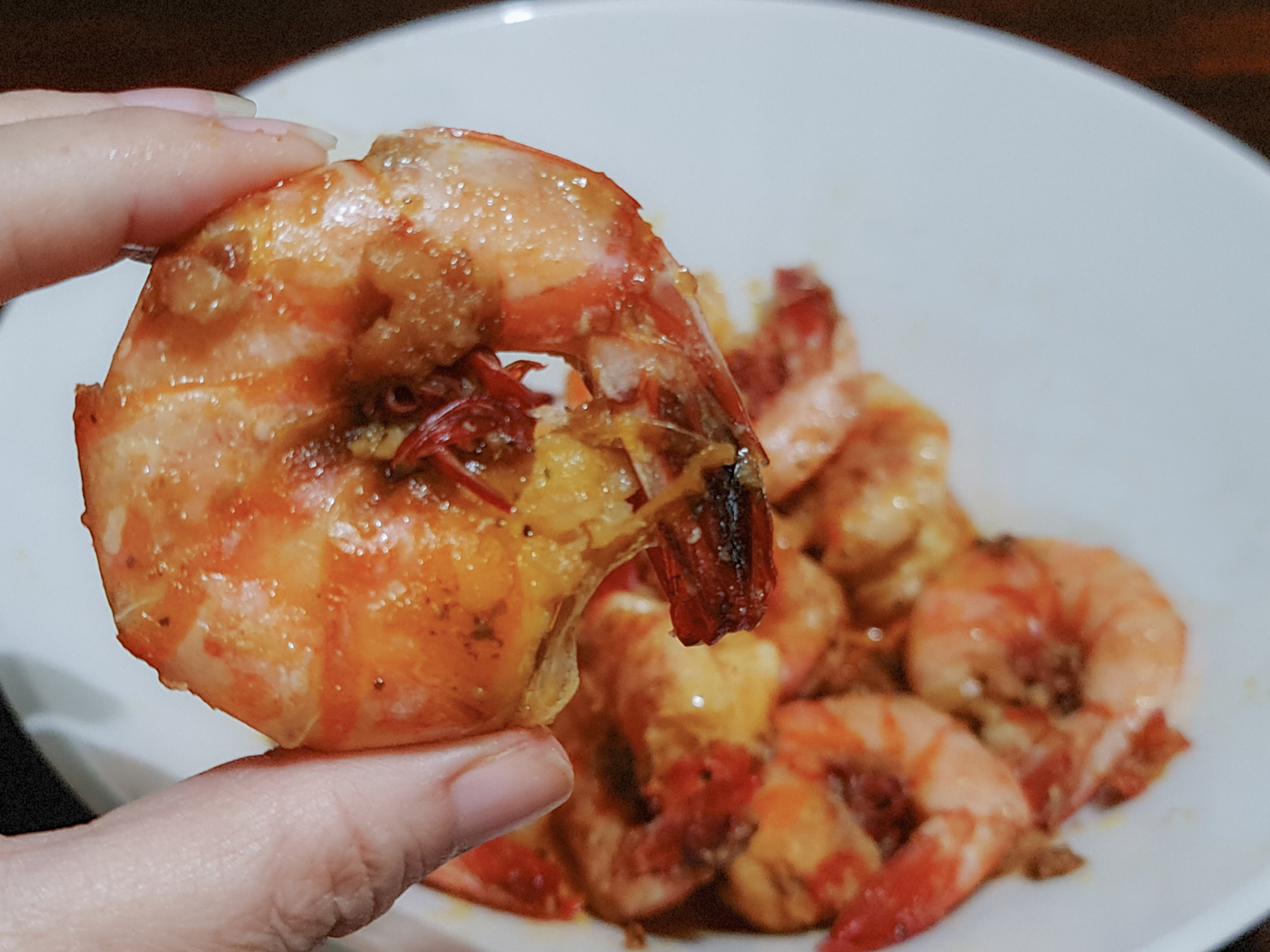 Close-up of a hand holding a cooked shrimp with more shrimp on a plate in the background