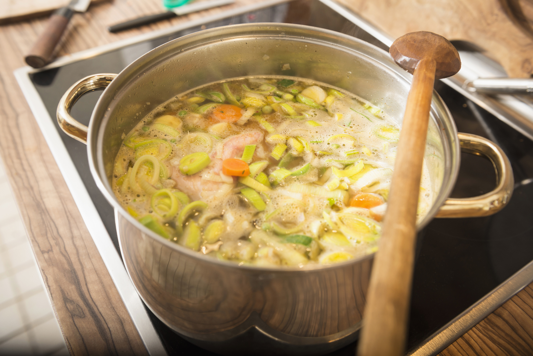A pot of soup with various vegetables and chicken on a stove, wooden spoon resting on the side