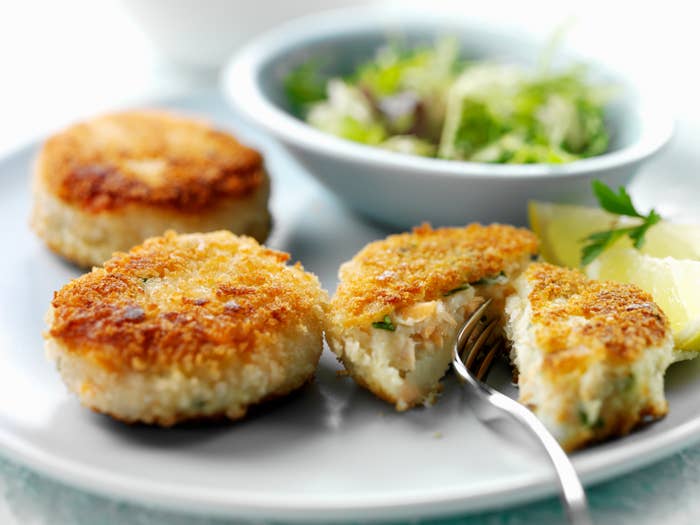 Three crab cakes on a plate with salad and a wedge of lemon, one is cut showing the inside