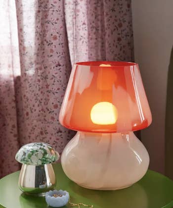 Table with a red lamp, a decorative mushroom, and jewelry beside curtains