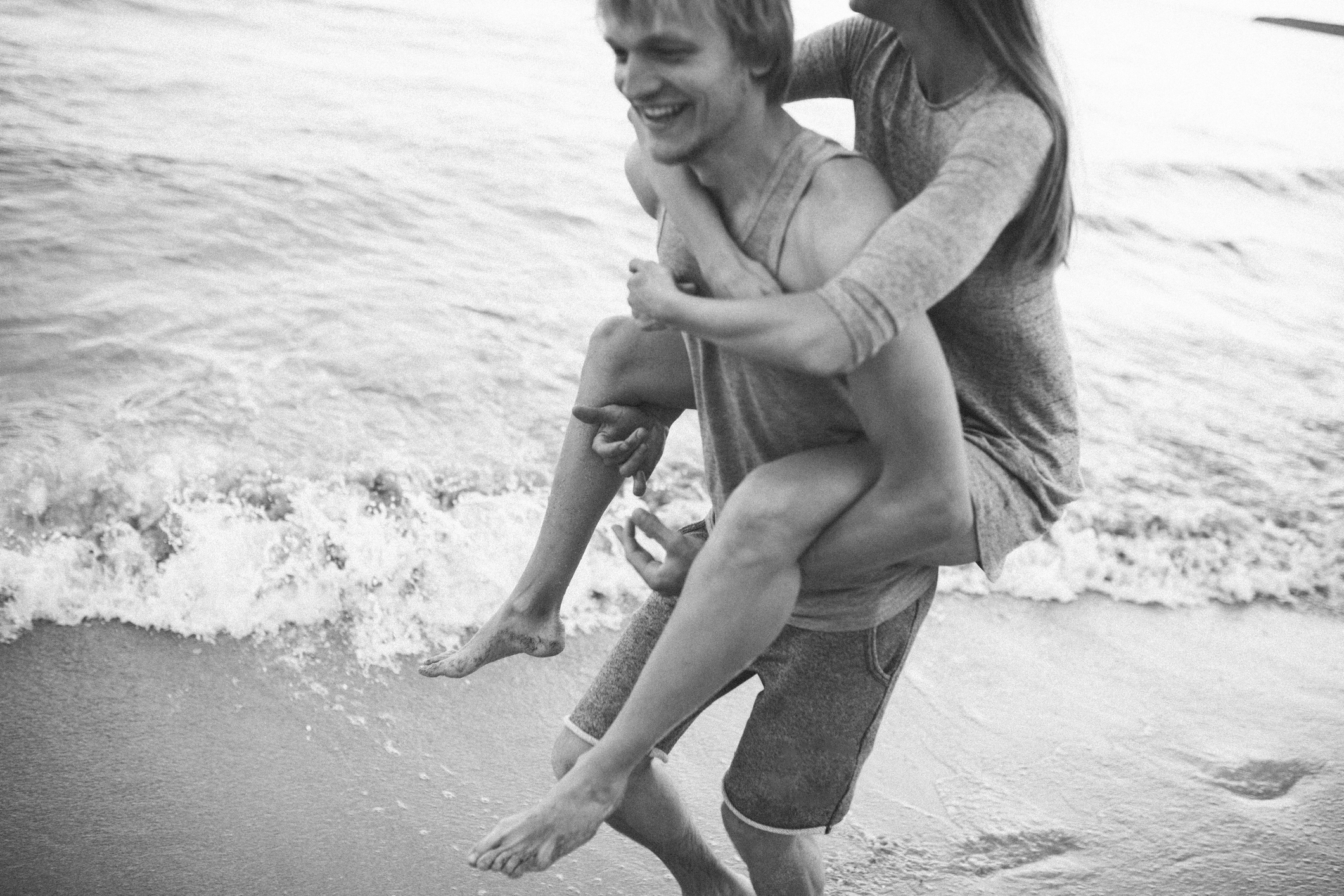 Man giving woman a piggyback ride on a beach, both smiling, waves in the background