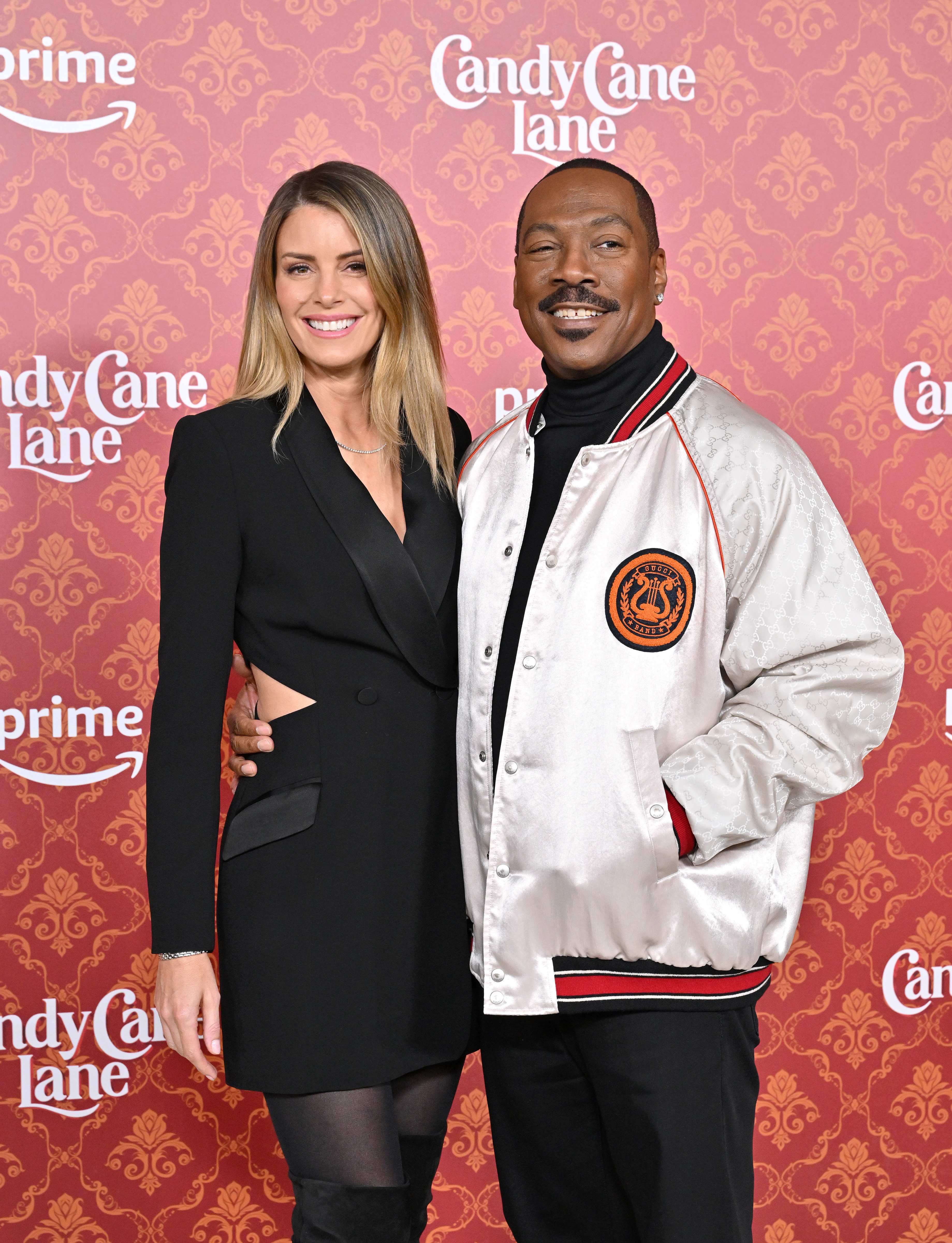 Two celebrities on a branded backdrop; woman in black dress, man in jacket with logo