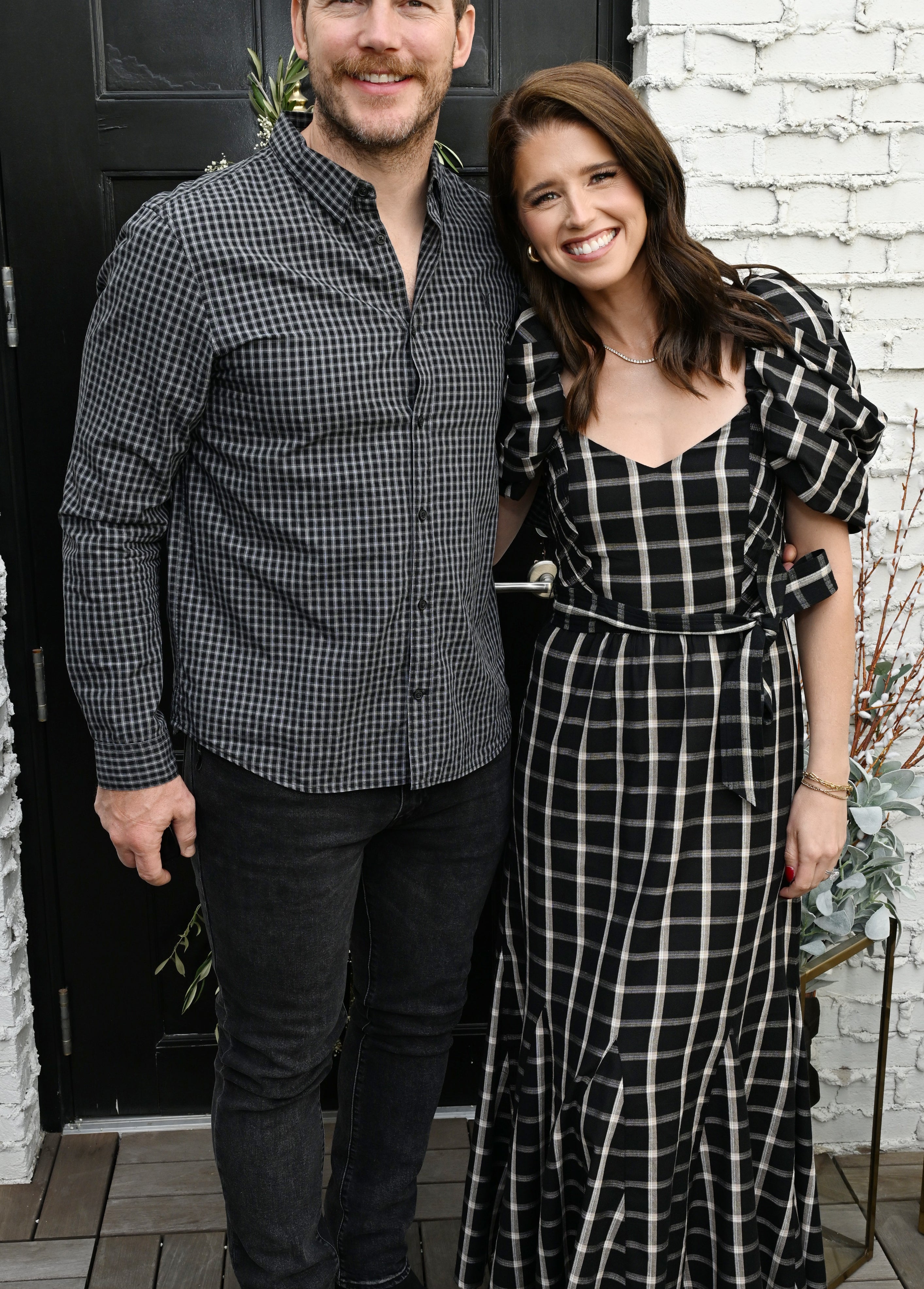 Two individuals standing together, one in a plaid dress with puff sleeves, the other in a checkered shirt and jeans