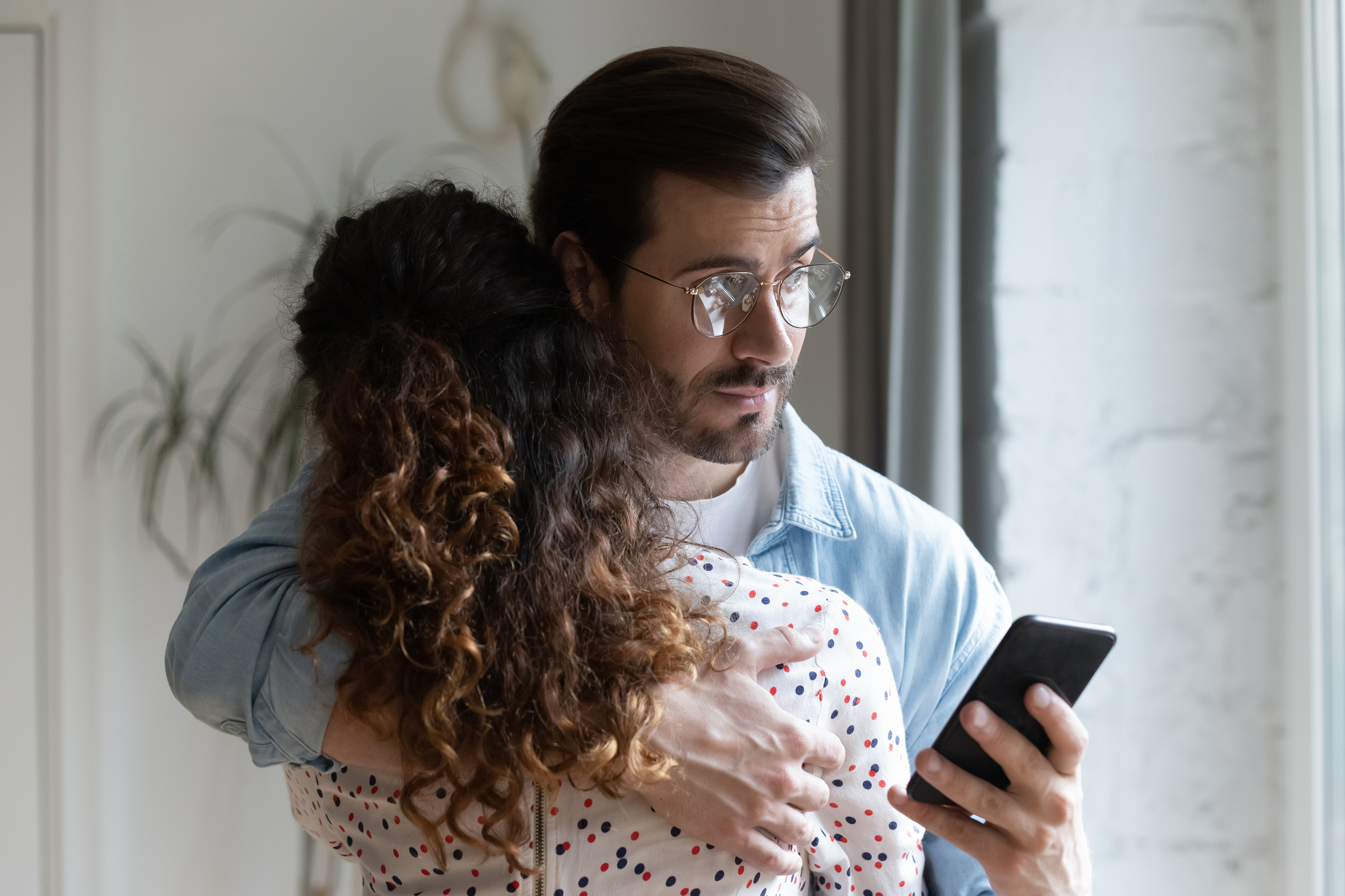 Man with glasses hugs a woman, both looking at his phone with concerned expressions