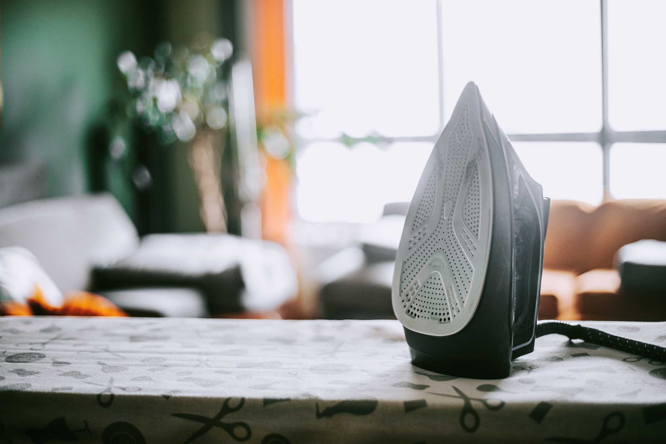 An iron placed upright on an ironing board with a blurred home background