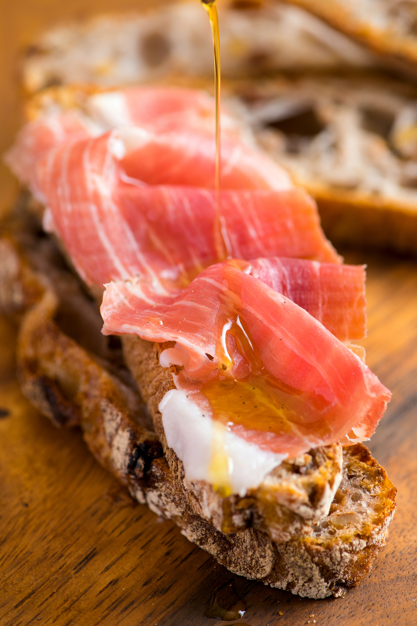 Honey drizzling on toast with prosciutto, next to sliced bread on a wooden board