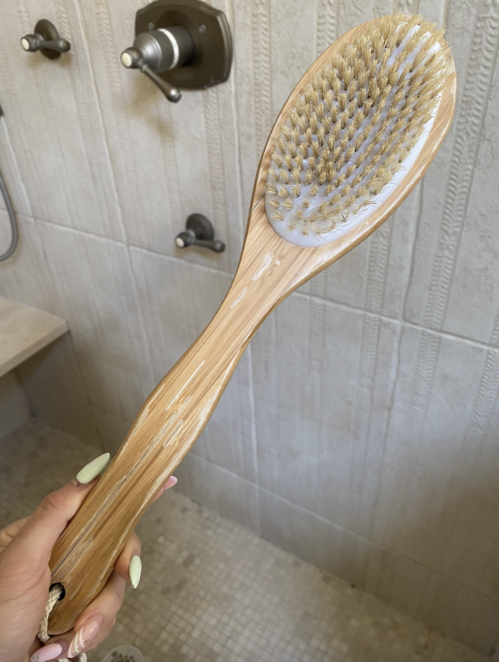 Hand holding a wooden bath brush with bristles, in front of a tiled shower wall