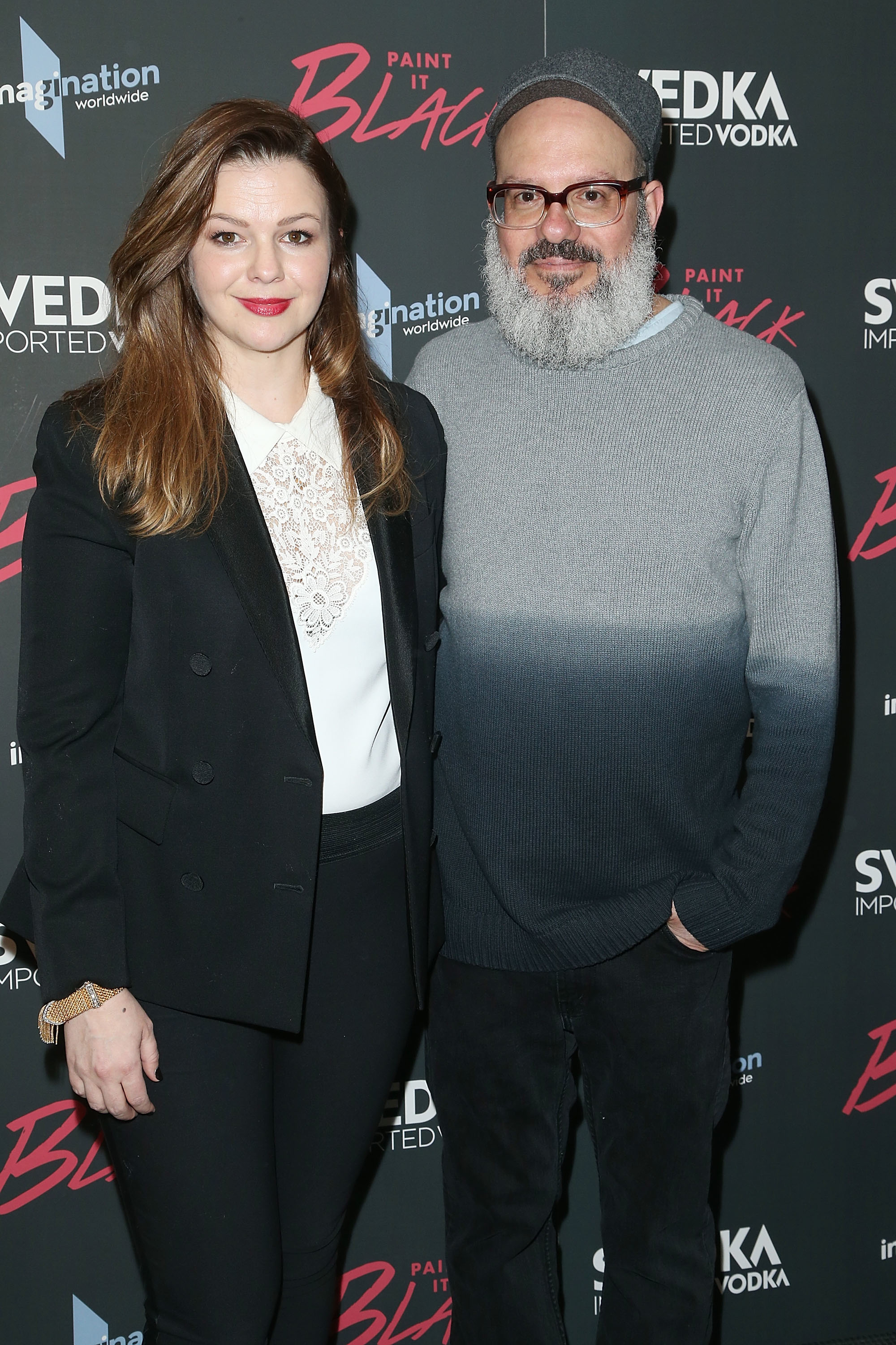 Two individuals posing together; one in a black blazer and white top, the other in a gray sweater. They are at an event with a branded backdrop