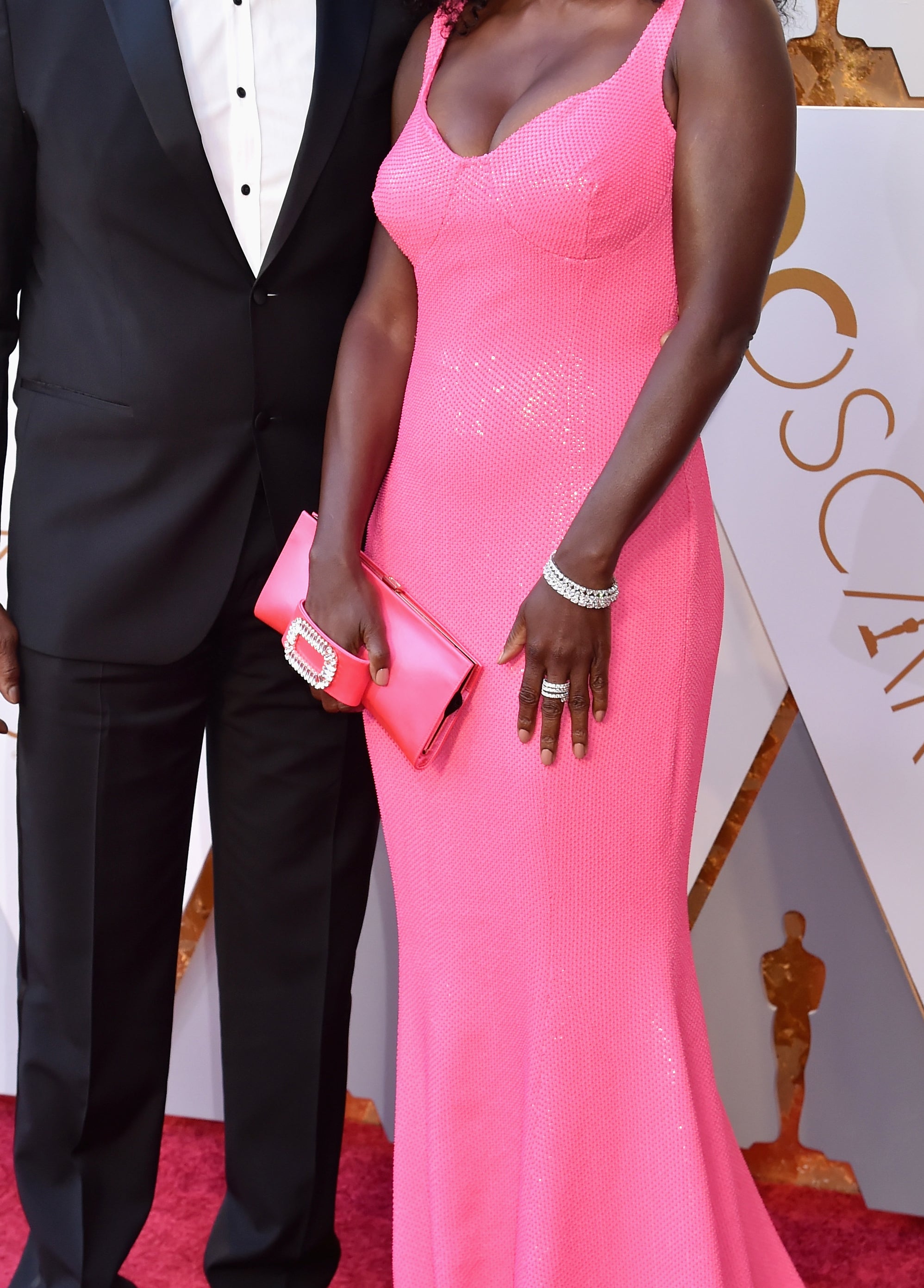 Two individuals posing at the Oscars, one in a black tuxedo, the other in a pink sleeveless gown with a clutch