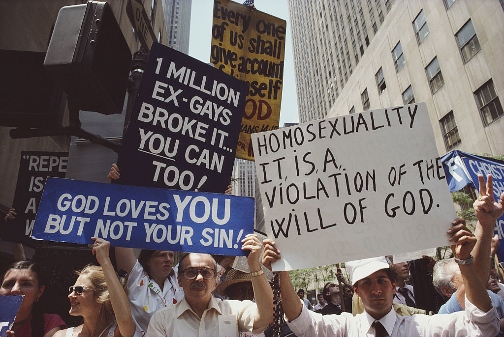 The image shows multiple people holding up protest signs with anti-gay messages