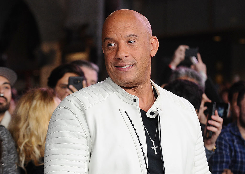 Vin Diesel wearing a white jacket and cross necklace, smiling at an event with crowd in the background