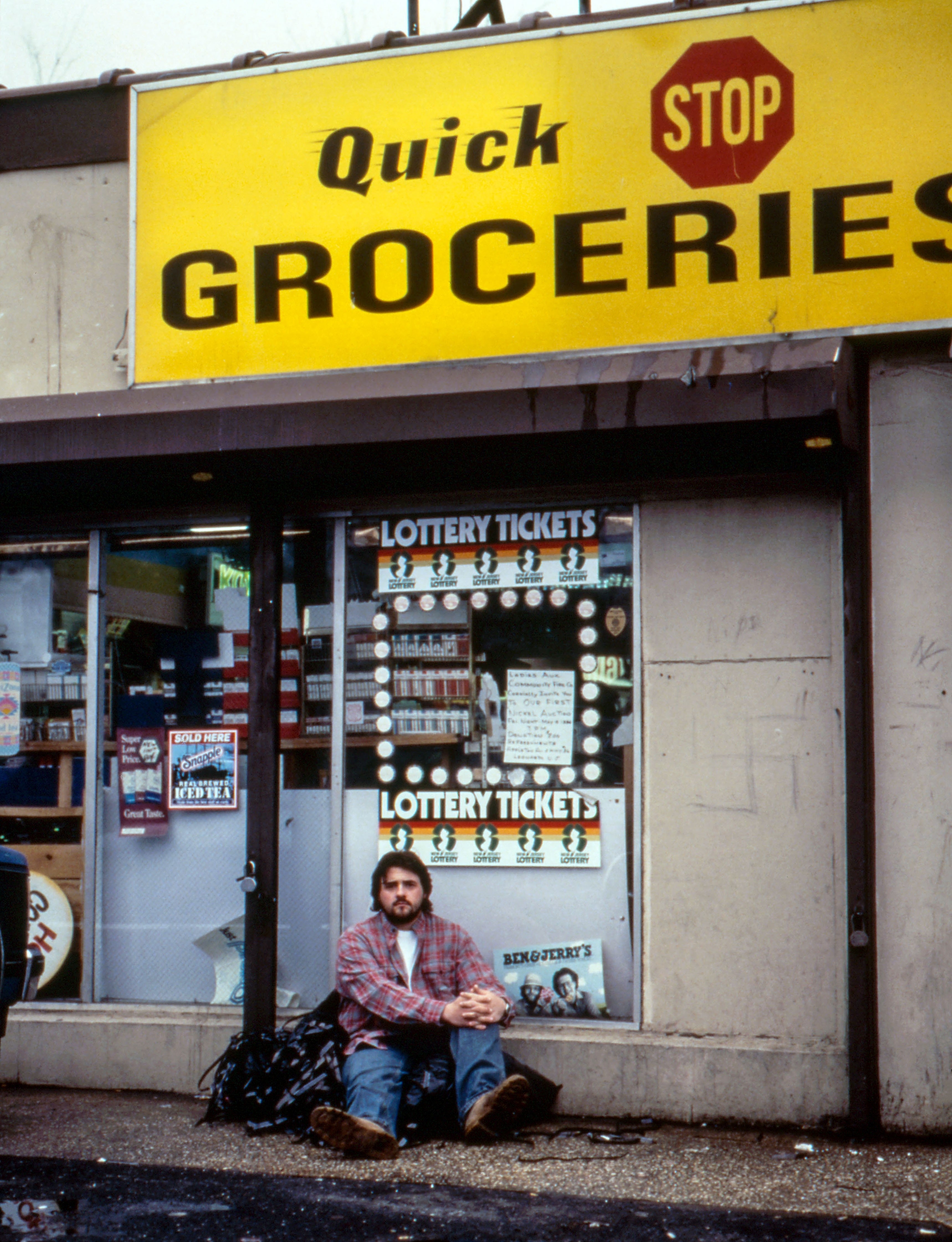 Kevin on location seated in front of a grocery store under a sign, with a reflective expression