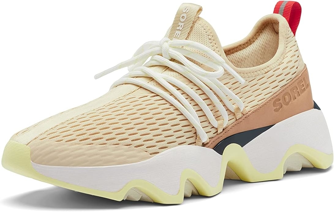 A modern athletic shoe with a mesh upper and chunky sole, featuring a beige and white design with a red pull tab
