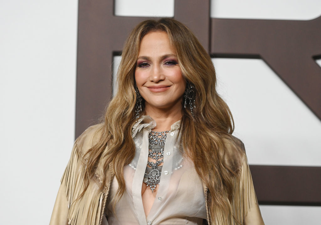 Jennifer Lopez is smiling, wearing a fringed jacket with embellished detailing and sheer blouse