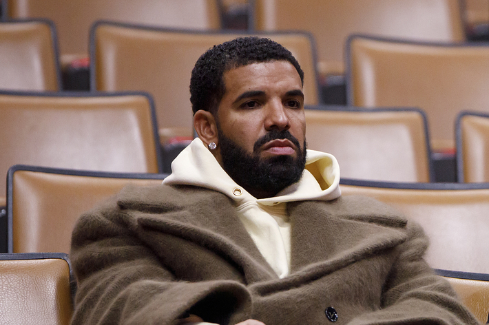 Drake sits alone in an auditorium wearing a large coat with a thoughtful expression