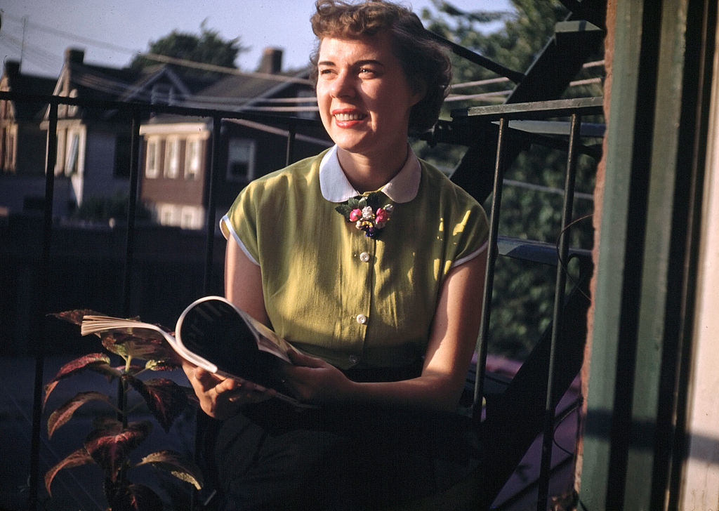 Woman in vintage attire smiling while sitting with book on balcony