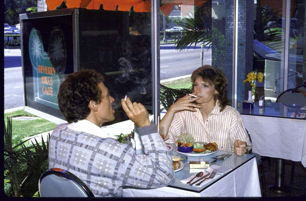 Two people conversing at a cafe table, one person smoking, with a visible dessert plate