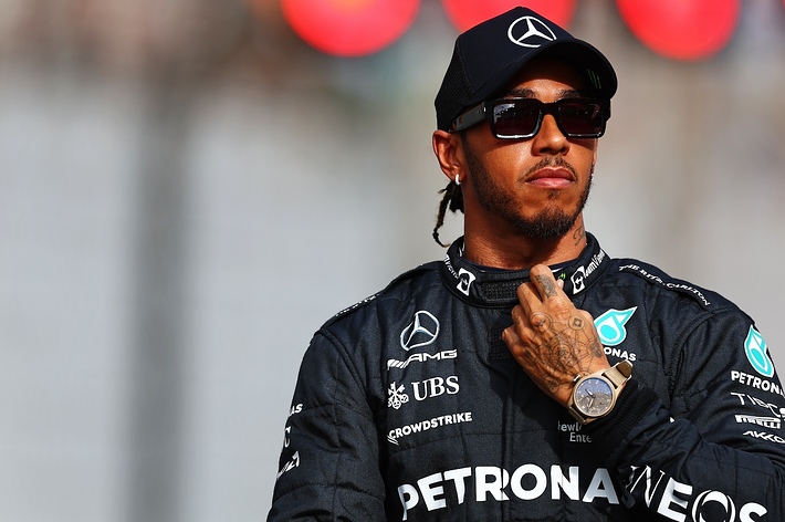 Lewis Hamilton in a racing suit, standing confidently with hand on heart