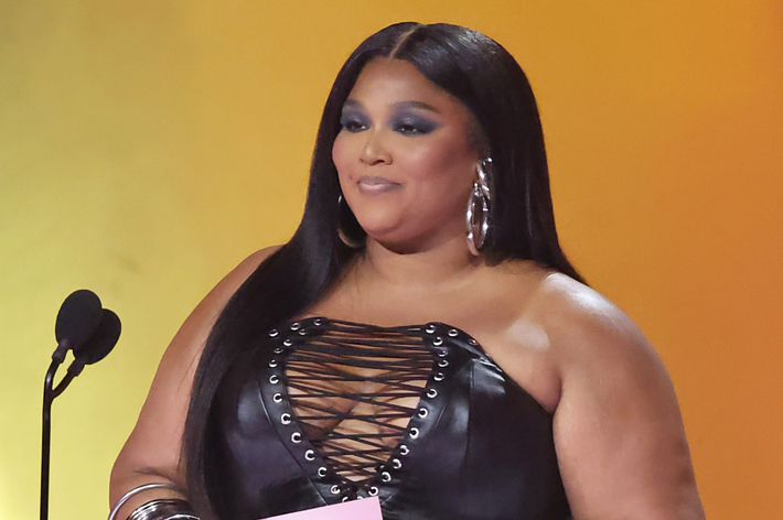 Lizzo stands at a podium with a microphone, wearing a laced-up outfit and earrings