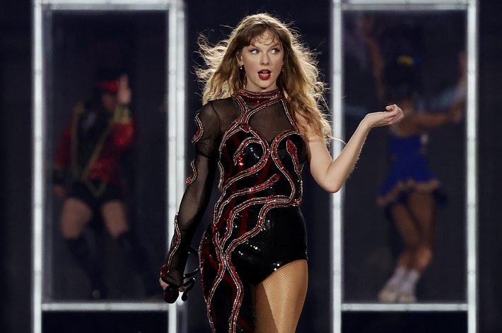 Taylor Swift performing on stage wearing a sparkling bodysuit with red accents and boots, with backup dancers in the background