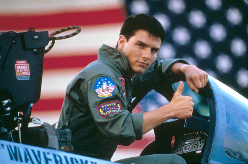 Tom Cruise as Maverick in a bomber jacket, giving a thumbs up beside a fighter jet