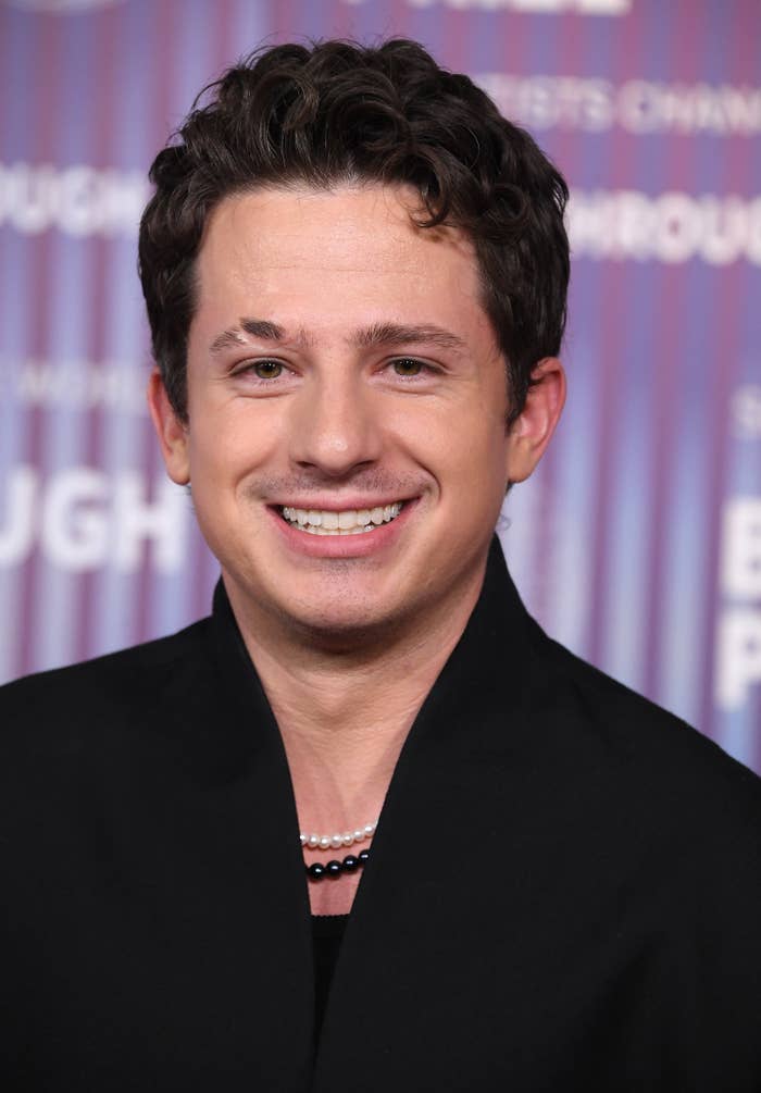 A closeup of Charlie Puth wearing a jacket and a necklace and smiling at an event