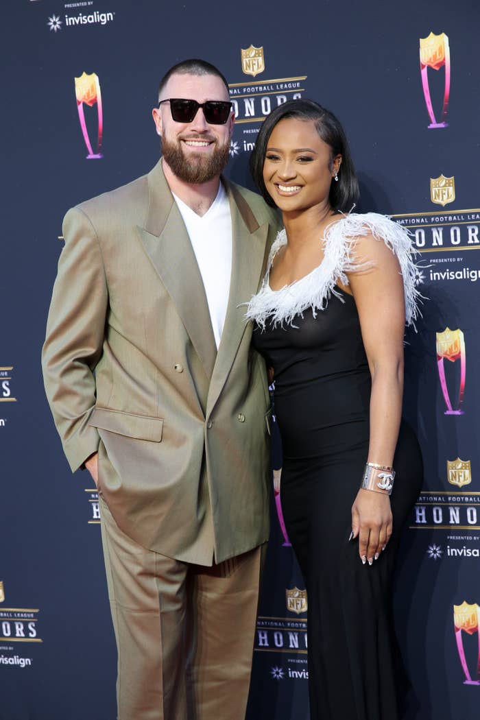 Travis Kelce in a suit and Kayla Nicole in a dress with feather details pose on the NFL Honors red carpet