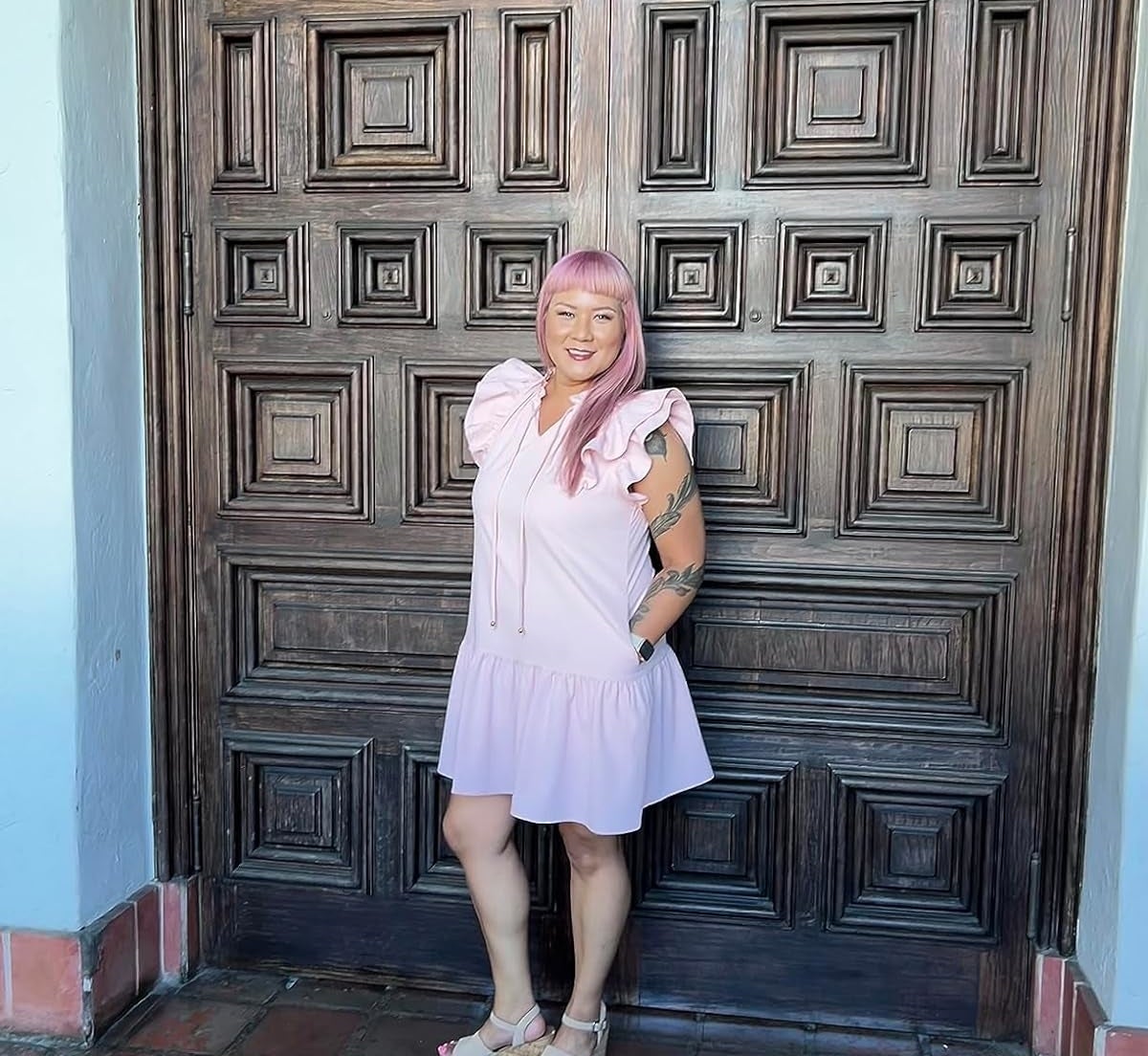 Person in a pink dress with frilled sleeves and sandals stands in front of a large wooden door